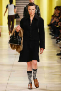 Model walking a runway in a black tailored outfit, carrying a fur stole and a glossy black handbag. She wears patterned socks with brown loafers.