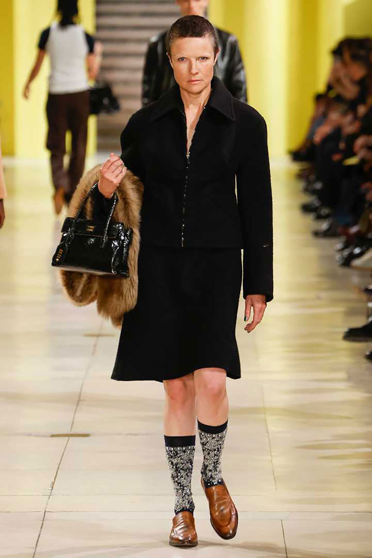Model walking a runway in a black tailored outfit, carrying a fur stole and a glossy black handbag. She wears patterned socks with brown loafers.