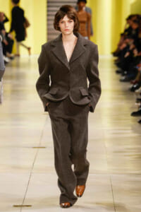 Model walks a fashion runway in a dark gray tailored suit with structured shoulders and a peplum-style jacket, matching wide-leg trousers, in a yellow-lit venue.