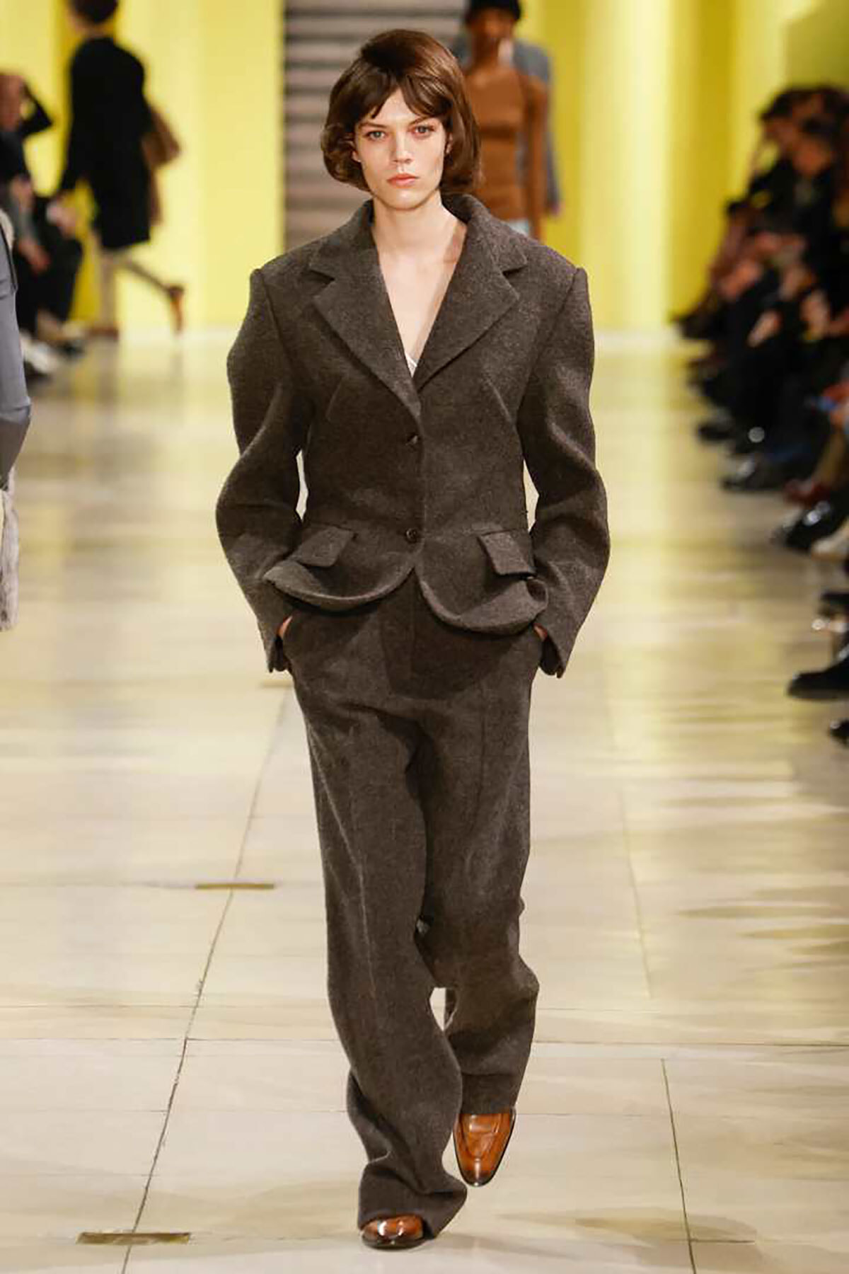 Model walks a fashion runway in a dark gray tailored suit with structured shoulders and a peplum-style jacket, matching wide-leg trousers, in a yellow-lit venue.