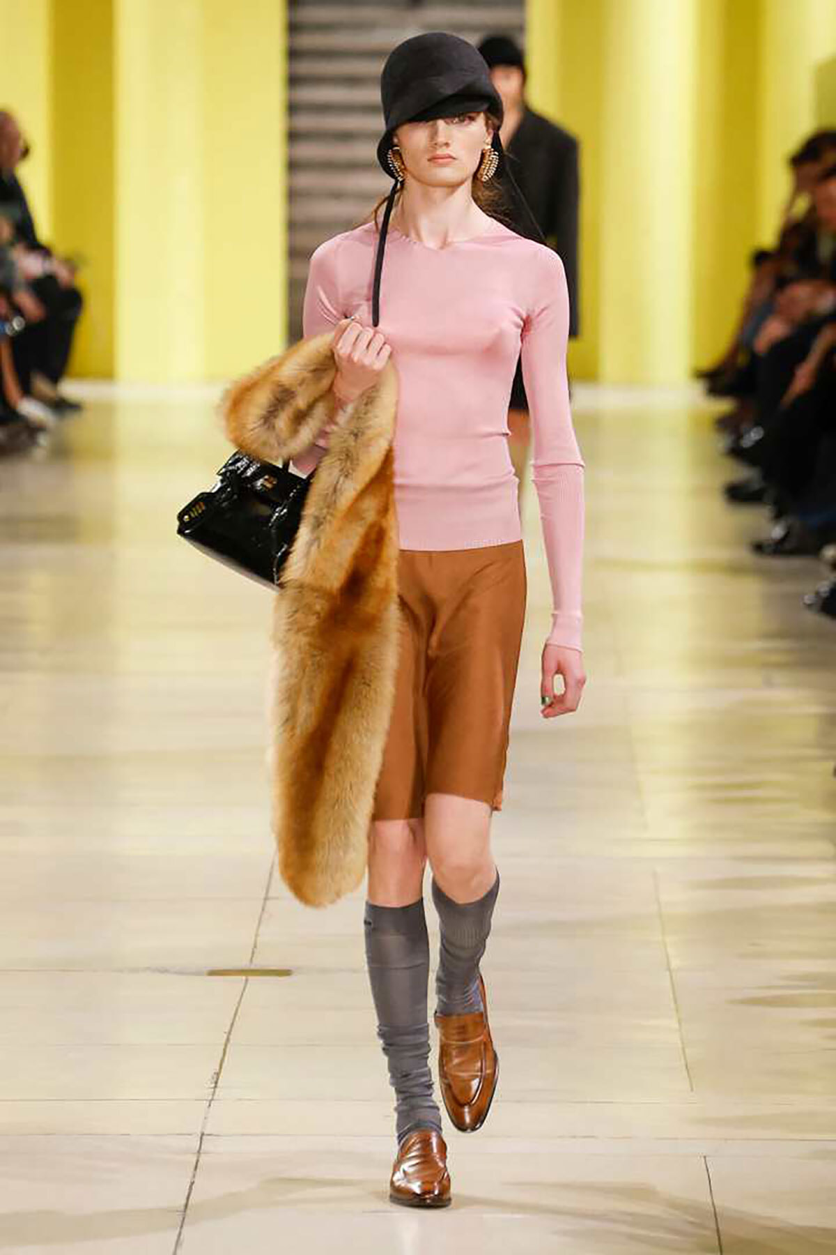Model walking a runway in a pink fitted long-sleeve top, camel shorts, gray leg warmers, brown loafers, a black hat, and a fur stole draped over her arm.
