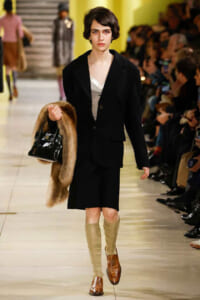 Runway model wears a black blazer and skirt with beige leg warmers, brown loafers, a fur stole, and a glossy black handbag as the audience watches.