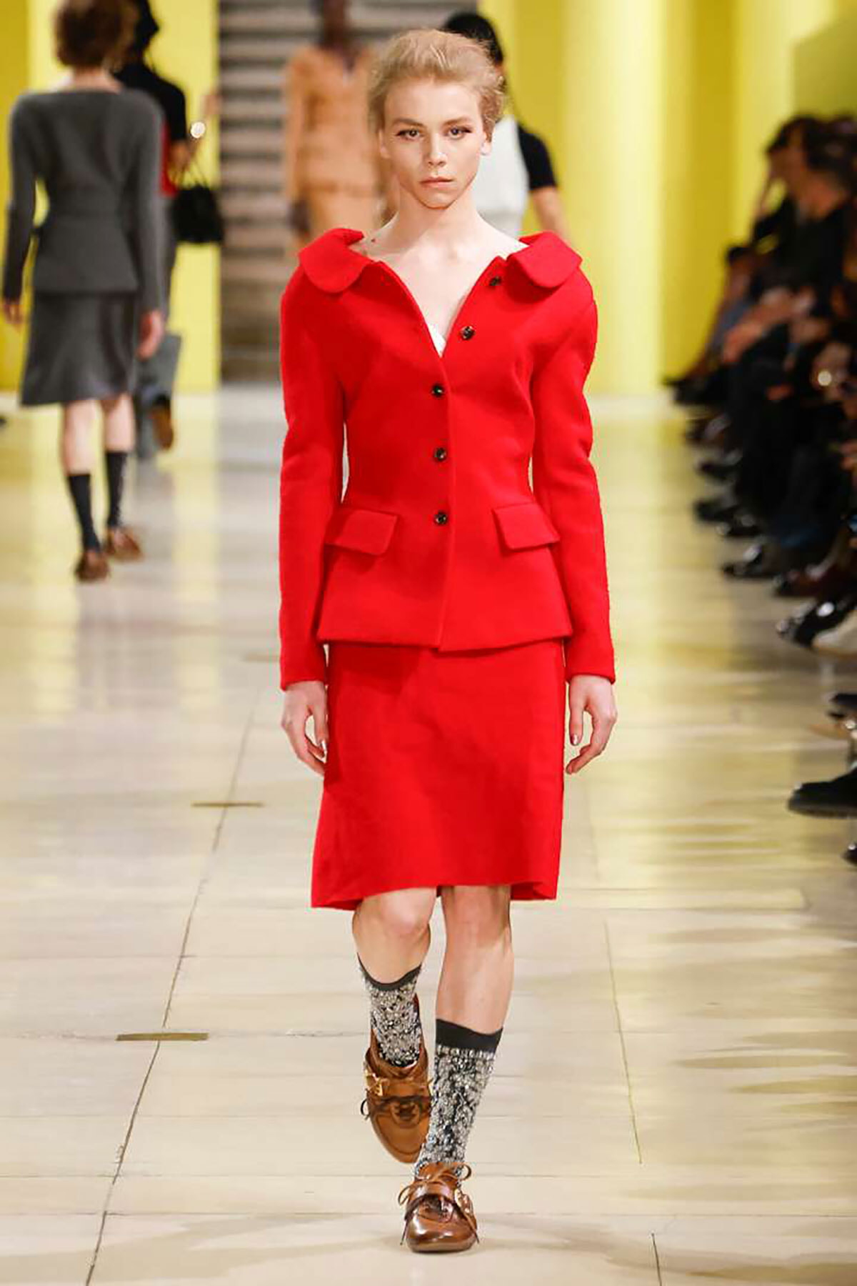 Model walks the runway in a red tailored suit featuring a double-breasted jacket and knee-length skirt on a fashion-show runway with a yellow backdrop.