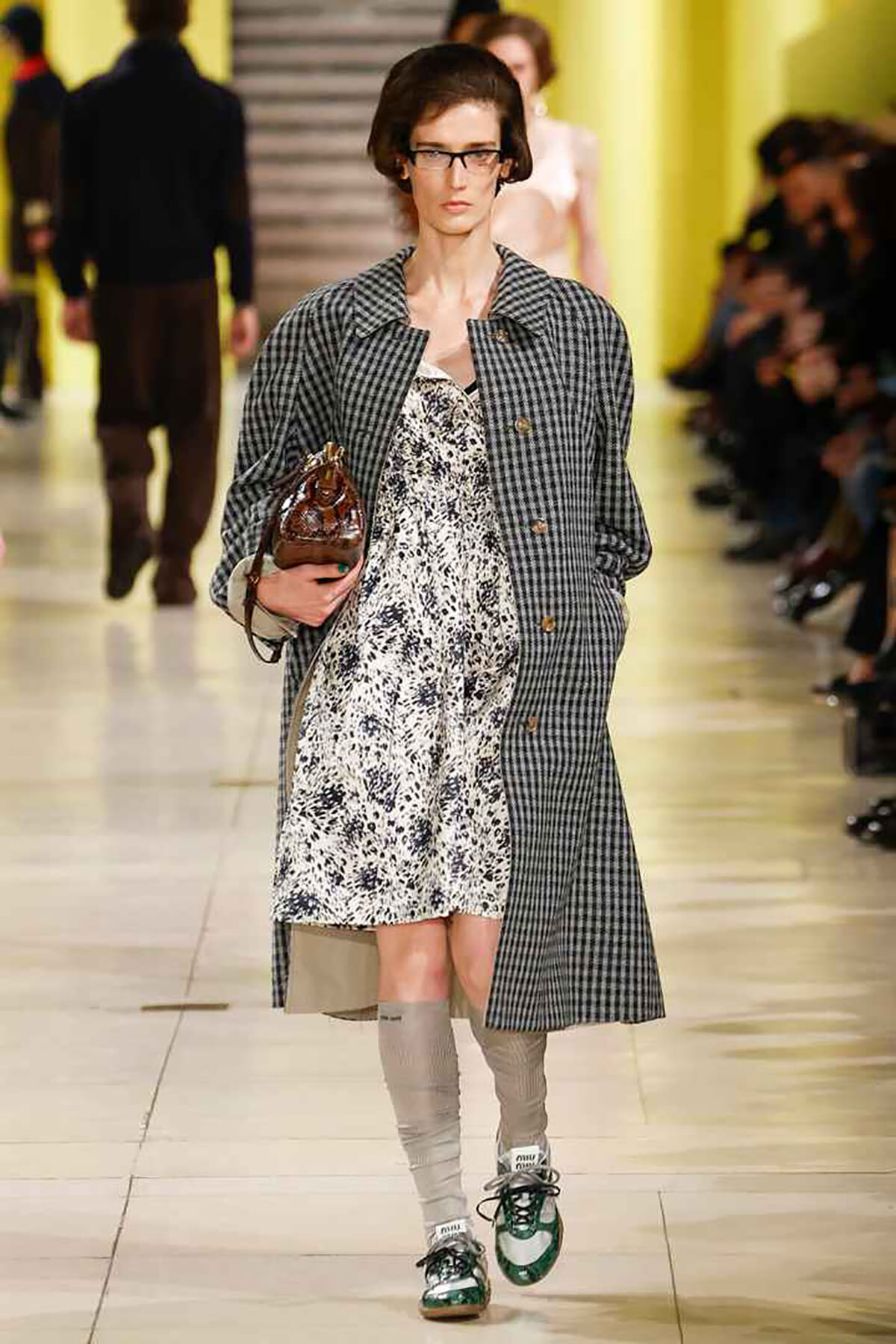 Model walks the runway in a gray checkered coat over a white floral dress, carrying a brown handbag, with gray knee-high socks and green-and-white sneakers.