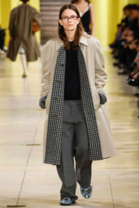 Model walking a fashion runway in a beige trench coat, black top, gray pants, and a long black-and-white checkered scarf.