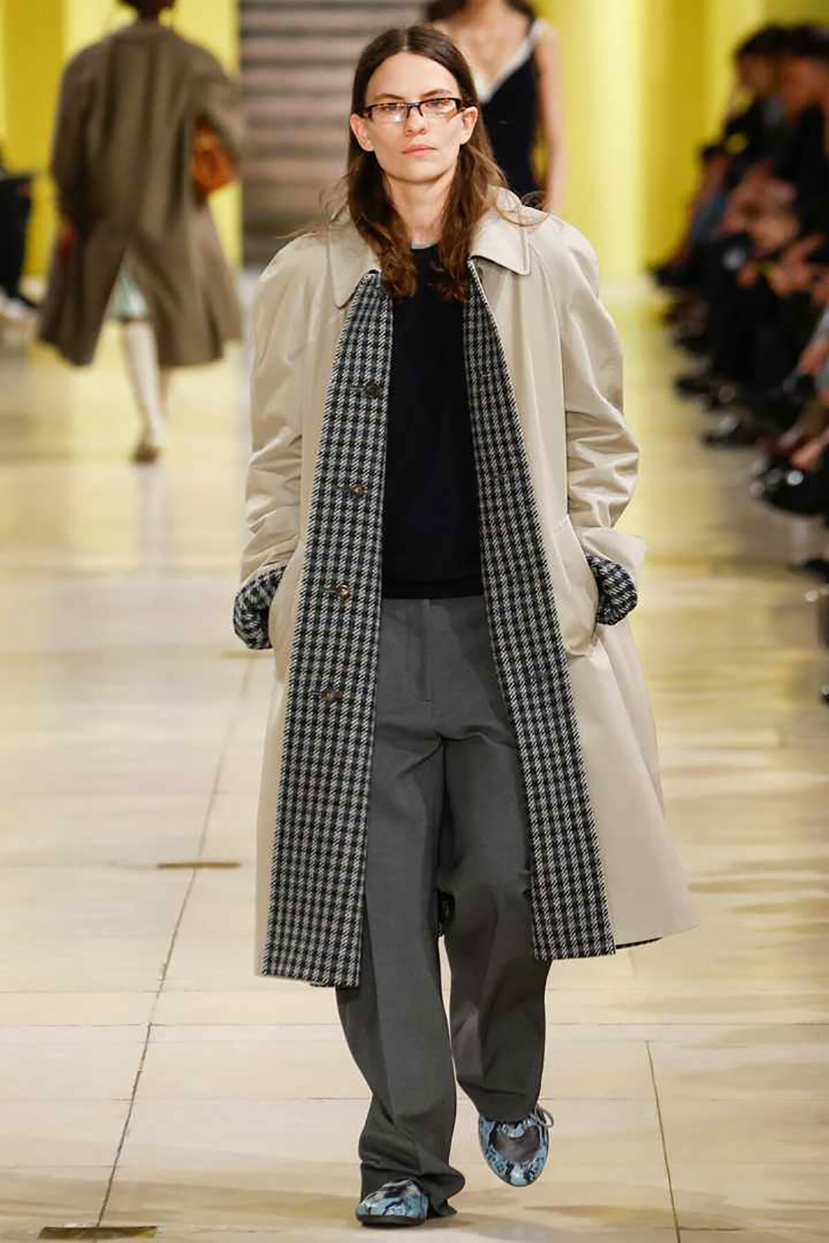 Model walking a fashion runway in a beige trench coat, black top, gray pants, and a long black-and-white checkered scarf.