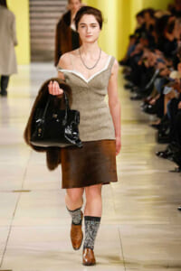 Model walks a runway in a cream knit top, brown leather skirt, fur stole, and a black handbag.