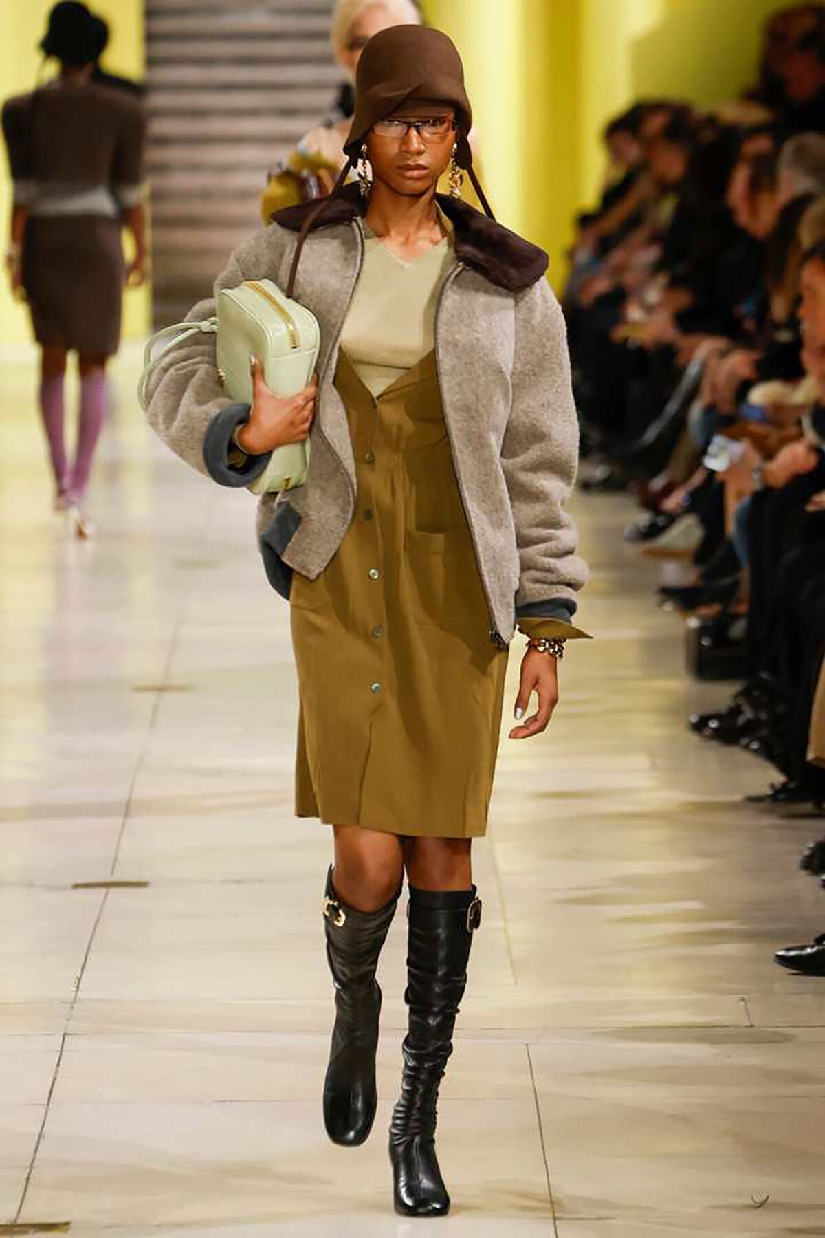 Model on runway wearing a gray shearling jacket over an olive dress, black knee-high boots, a brown hat, and a mint handbag.