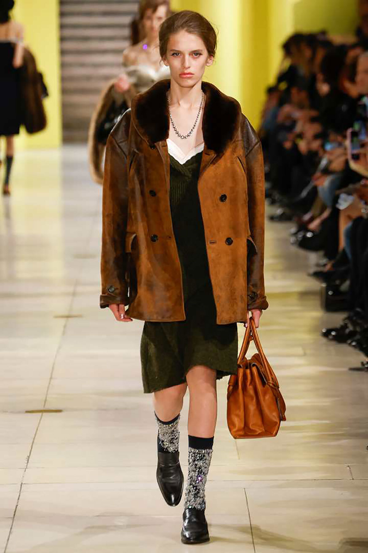 Female model walks the runway in a brown suede coat with fur collar over a green dress, carrying a brown handbag.
