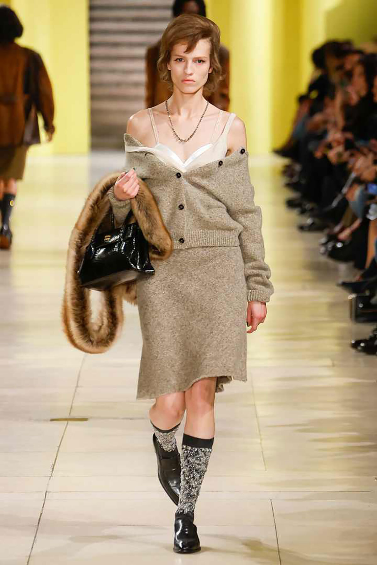 Model walks a runway in a beige knit cardigan draped off shoulders over a white bralette, grey midi skirt, fur stole, and black patent bag; sparkly socks and ankle boots.
