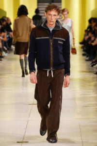 Male model walking a fashion runway in a navy-brown colorblock zip jacket and brown drawstring pants on a beige catwalk.