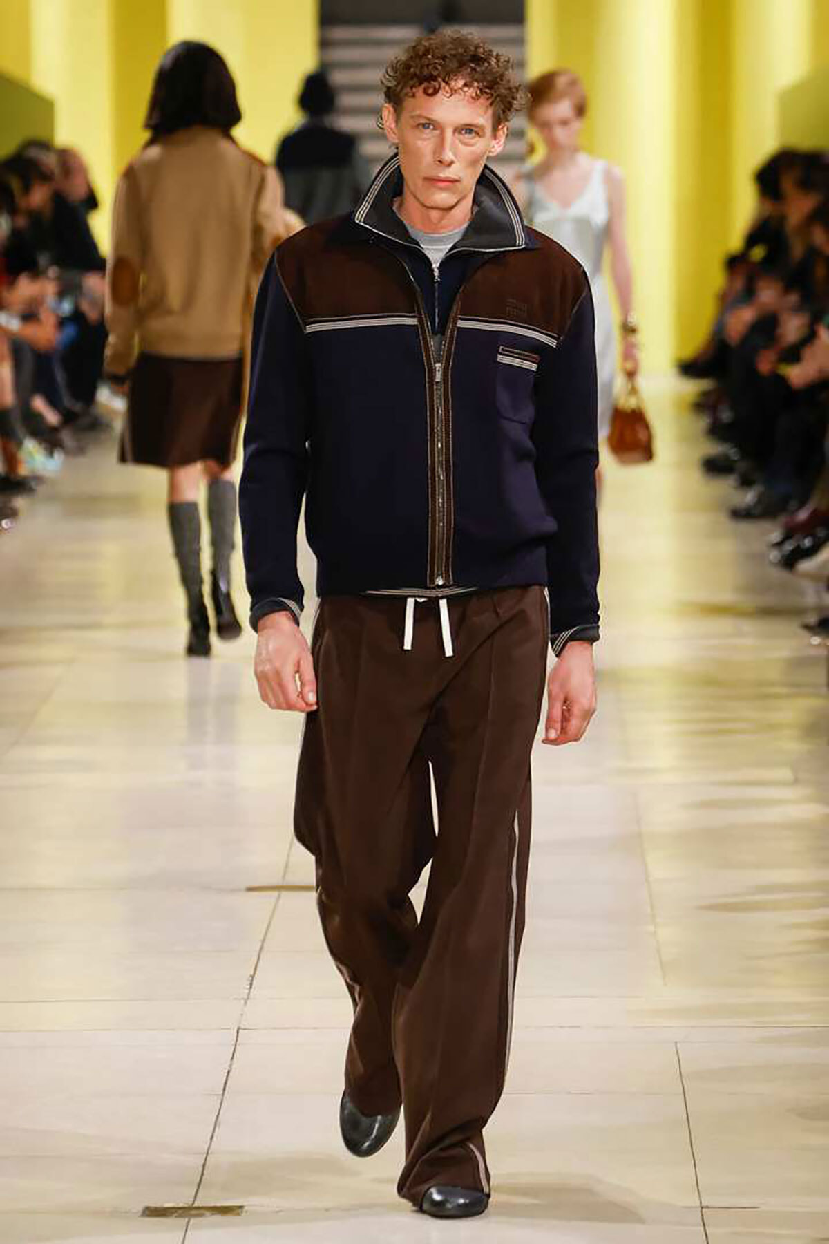 Male model walking a fashion runway in a navy-brown colorblock zip jacket and brown drawstring pants on a beige catwalk.
