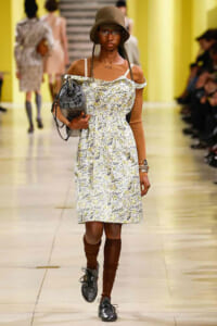 Model walks the runway in a floral off-shoulder dress with a brown hat, glasses, and a gray handbag.