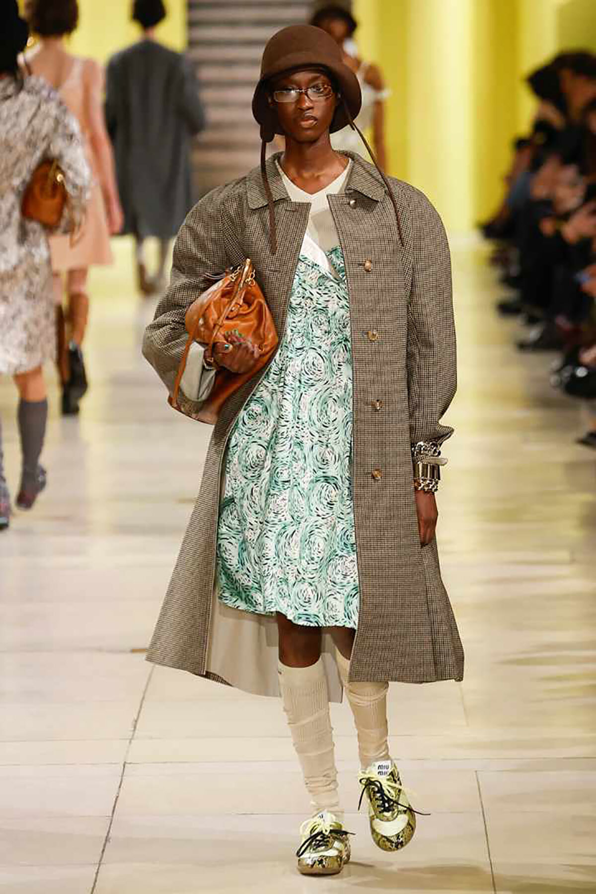 Model on a runway wearing a gray checkered trench over a turquoise floral dress, with a brown bucket hat and cream leg warmers, gold sneakers, and a brown bag.