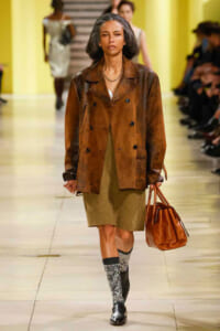 Model walking a runway in a brown leather double-breasted coat over an olive dress, carrying a tan leather tote and wearing glittery socks with black ankle boots.