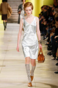 Model walking runway in a silver satin slip dress, tan handbag, gray knee-high socks, and brown shoes, with bold gold earrings and bracelets.