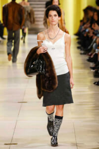 Model struts down a fashion show runway in a white camisole, gray skirt, patterned knee-high socks, black shoes, carrying a brown fur stole and a glossy black handbag, with a yellow backdrop in the background.