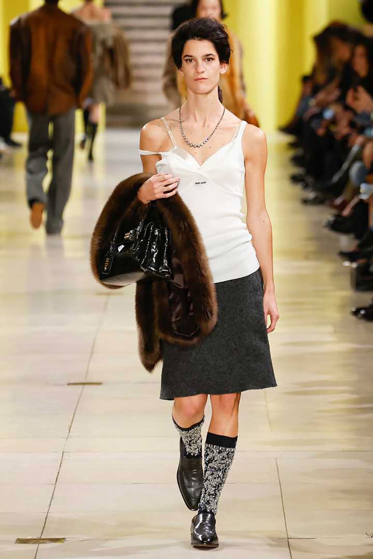 Model struts down a fashion show runway in a white camisole, gray skirt, patterned knee-high socks, black shoes, carrying a brown fur stole and a glossy black handbag, with a yellow backdrop in the background.