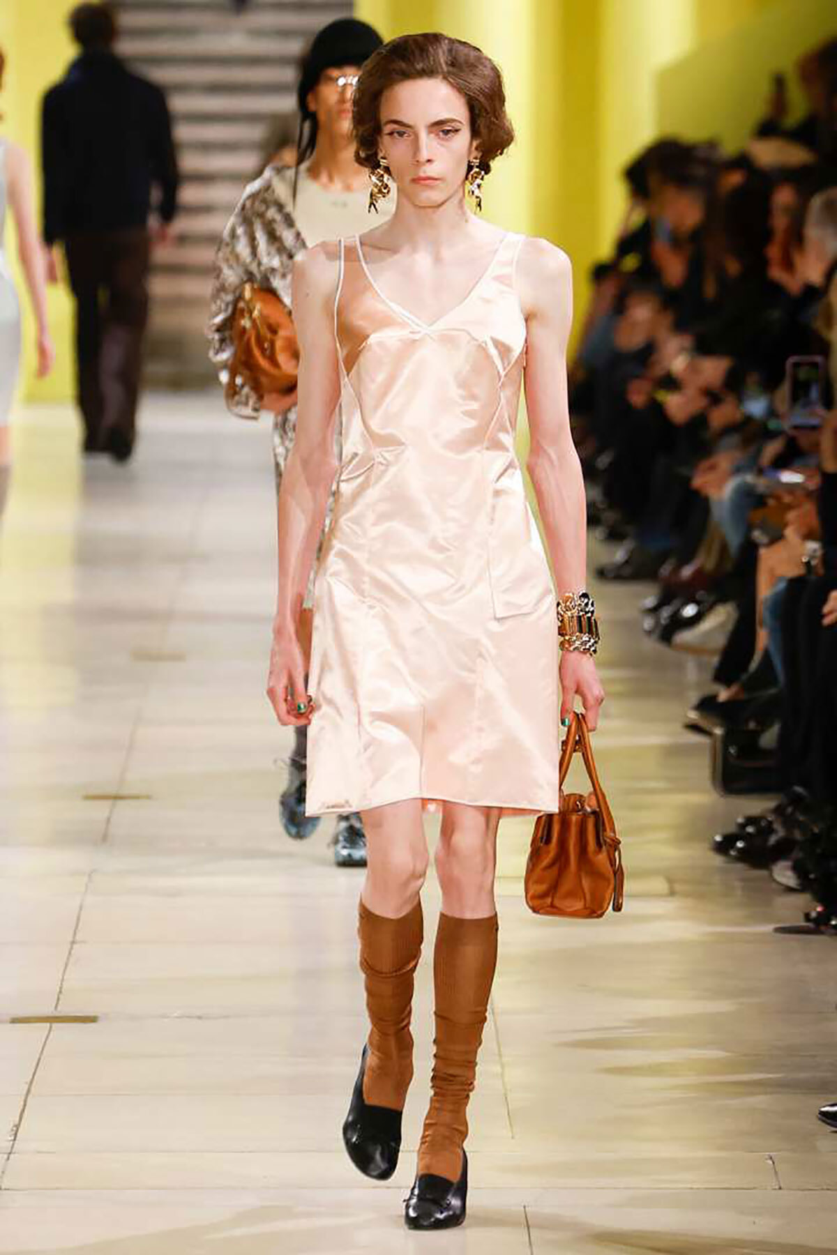 Model walks runway in pale pink satin slip dress, brown knee-high boots, and a brown handbag with gold jewelry.
