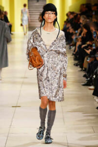 Model walking a runway in a patterned oversized coat, black hat, glasses, and gray leg warmers, carrying a brown shoulder bag.
