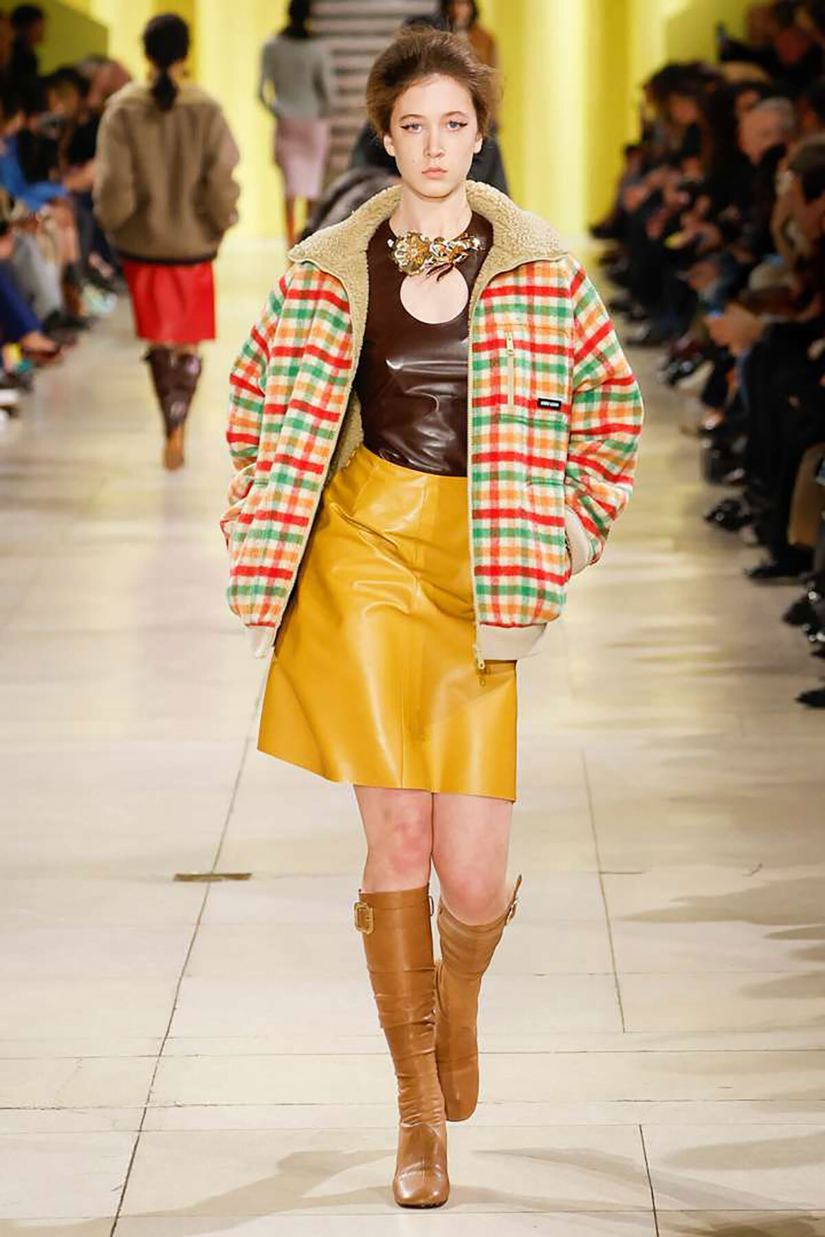 Model walking a fashion runway in a plaid bomber jacket, mustard leather skirt, brown top, knee-high tan boots, and statement necklace.