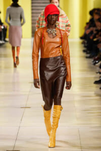 Model walking a fashion runway in a caramel-orange leather top, dark brown leather skirt, bright yellow knee-high boots, and a red hat with bold jewelry.
