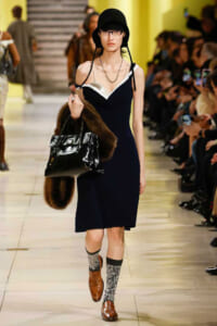 Runway model wearing a black knee-length dress, black hat, glasses, layered necklaces, carrying a fur stole and a glossy black handbag.