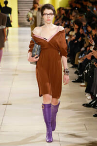 Model walking a runway in a brown off-shoulder dress with a pink neckline, purple knee-high boots, and a silver clutch.