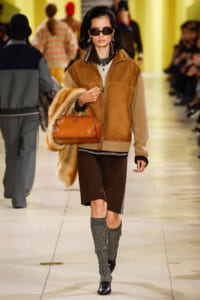 Model on a runway wearing a tan shearling jacket, brown shorts, gray knit knee-high boots, sunglasses, carrying a fur coat and tan leather handbag.