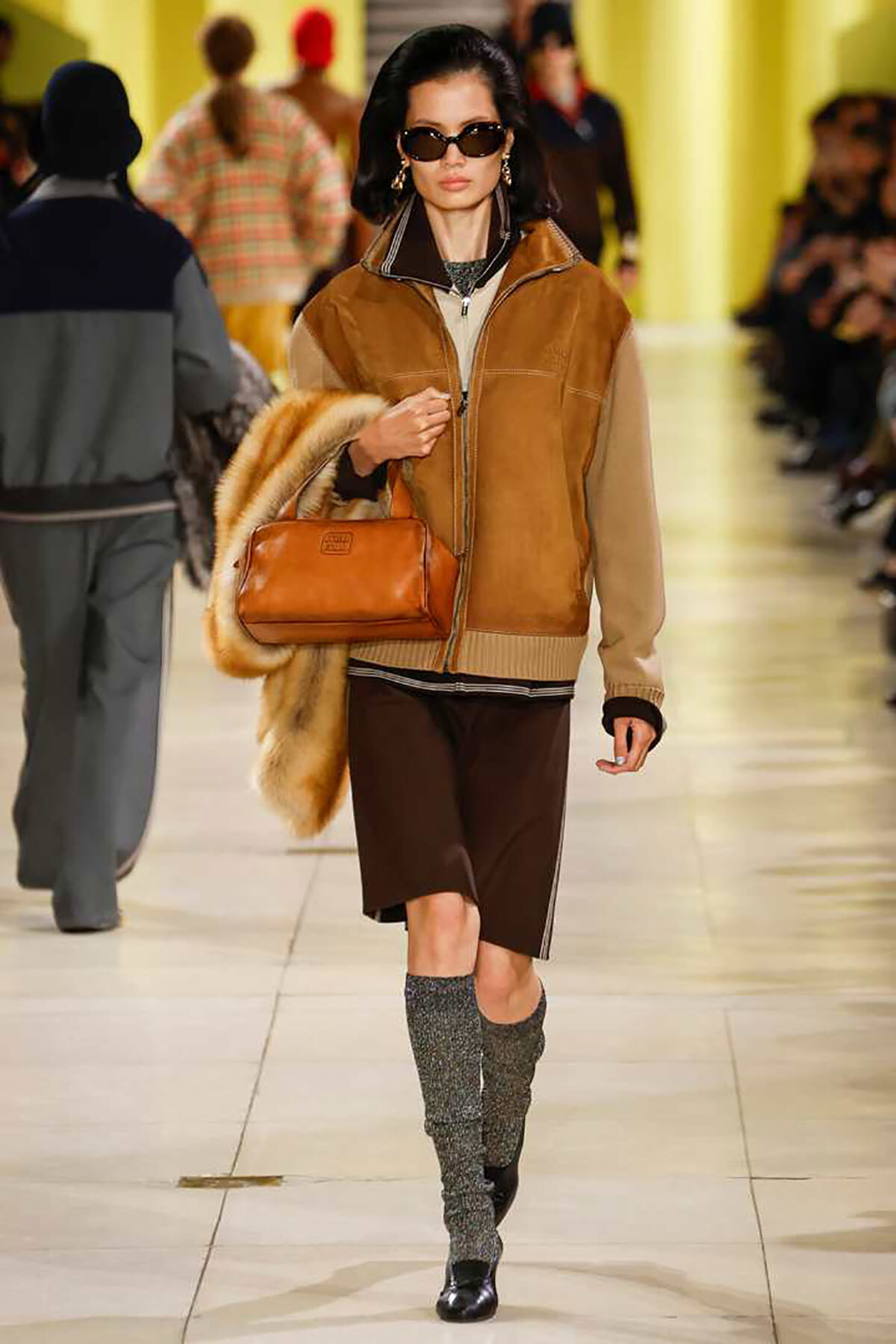 Model on a runway wearing a tan shearling jacket, brown shorts, gray knit knee-high boots, sunglasses, carrying a fur coat and tan leather handbag.