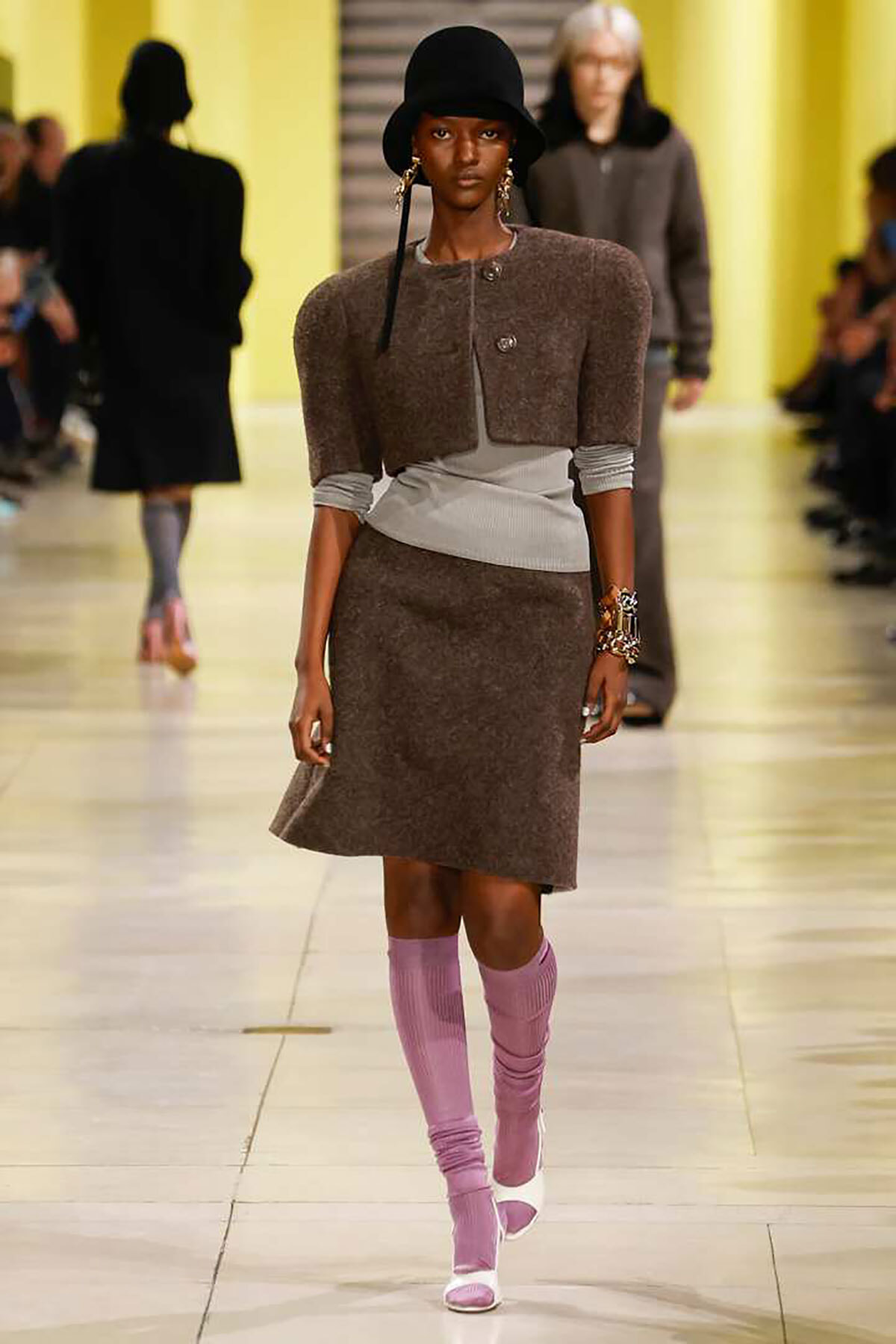 Model walks a fashion runway in a brown textured dress with a light gray wrap, black hat, and pink knee‑high socks.