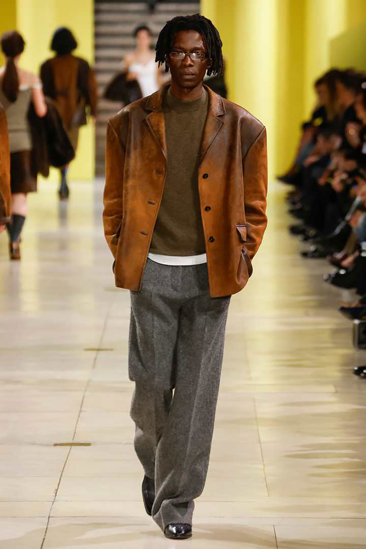 Male model on a fashion runway wearing a brown leather blazer, olive sweater, and gray wide-leg trousers.