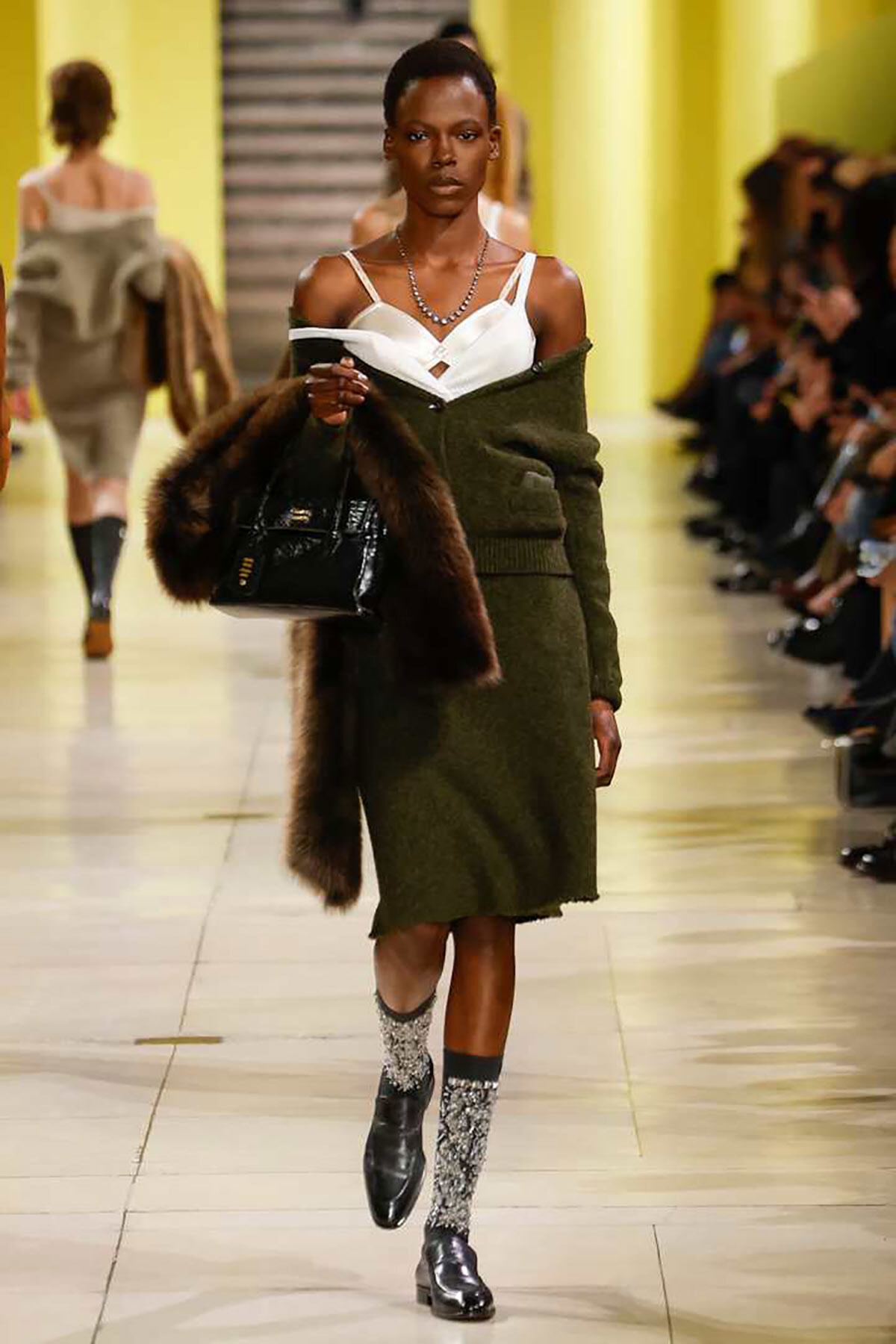 Model walking a fashion runway in an olive-green coat over a white top, carrying a fur stole and a black handbag, with glittery silver socks and black boots.