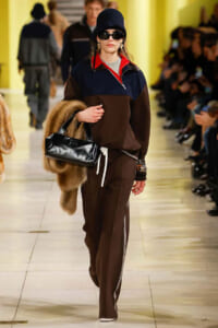 Model walks the runway in a color-block navy/brown pullover, matching brown pants with white side stripes, black hat and sunglasses, carrying a black patent handbag and a fur stole.