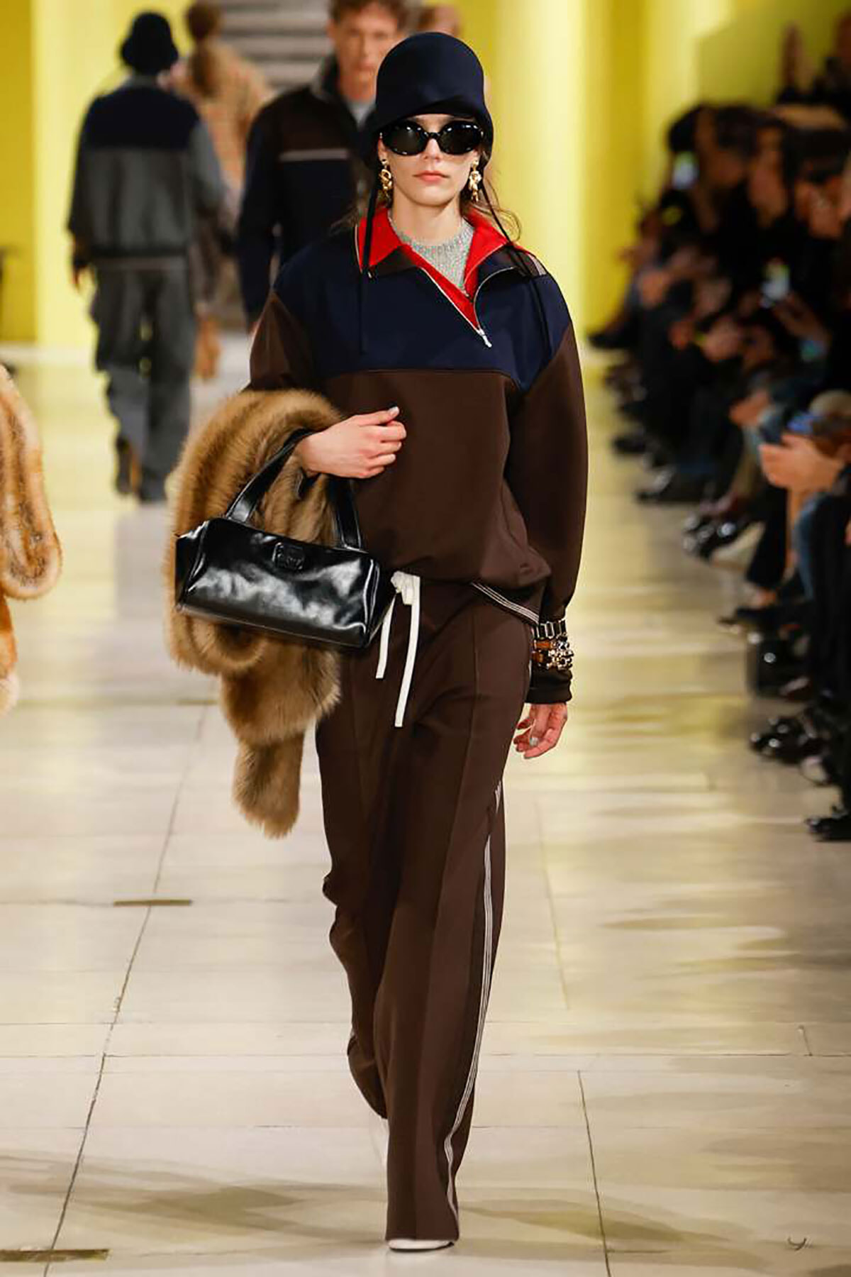 Model walks the runway in a color-block navy/brown pullover, matching brown pants with white side stripes, black hat and sunglasses, carrying a black patent handbag and a fur stole.