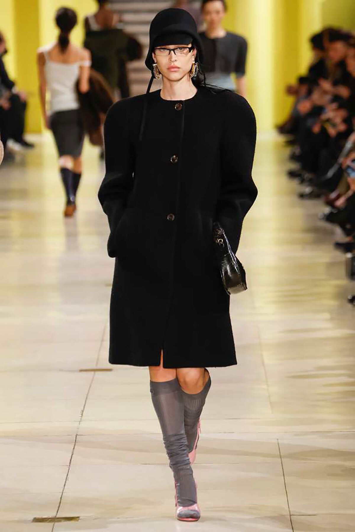 Model walking a fashion runway in a long black coat with a matching black hat, glasses, and bold hoop earrings, carrying a small black purse.