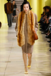 Model walks a runway wearing a beige textured coat with a long fur stole and camel boots on the catwalk.