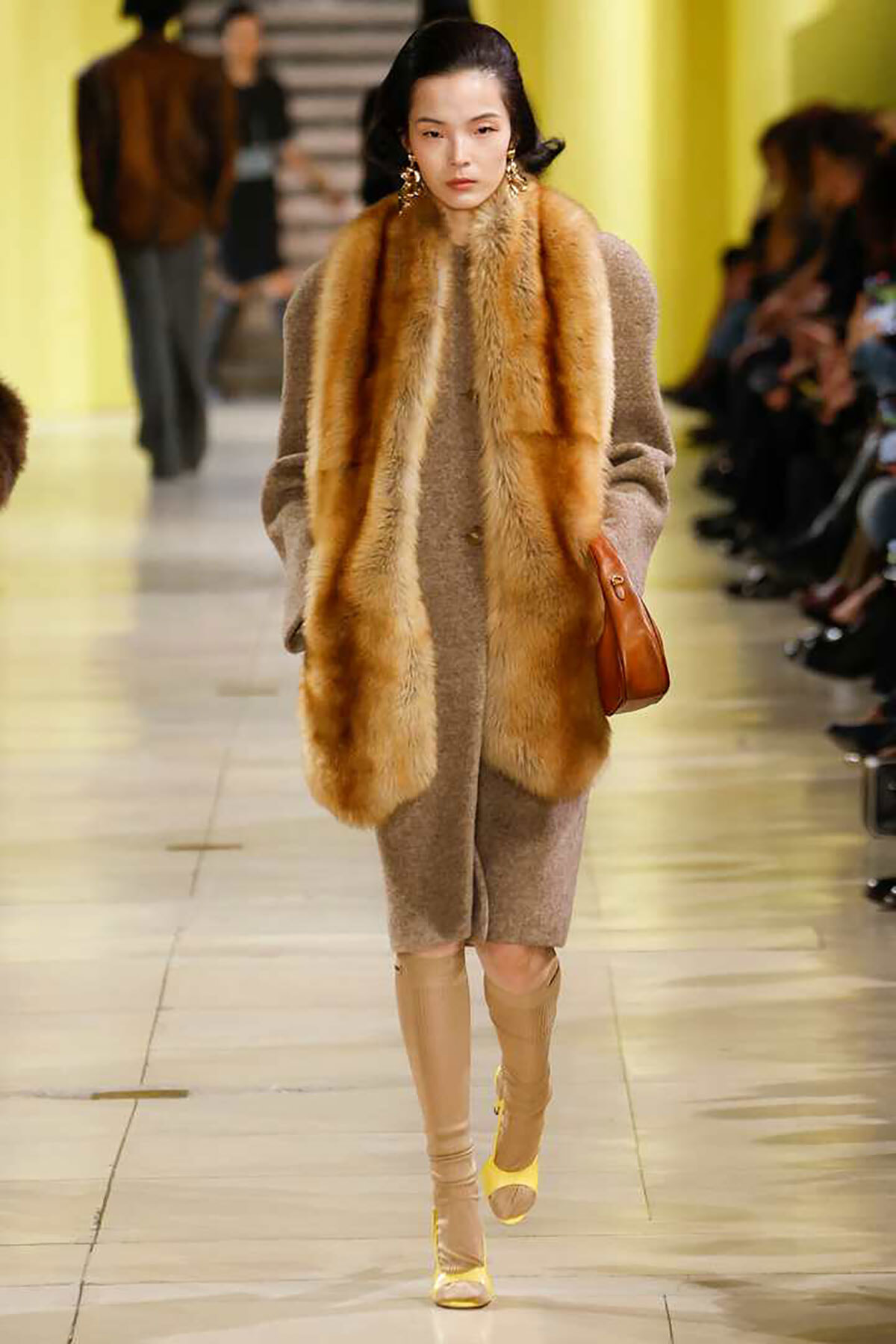 Model walks a runway wearing a beige textured coat with a long fur stole and camel boots on the catwalk.