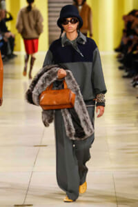 Model walking the runway in a color-block oversized gray and navy outfit, black hat, sunglasses, and yellow shoes, carrying a brown handbag and fur stole.