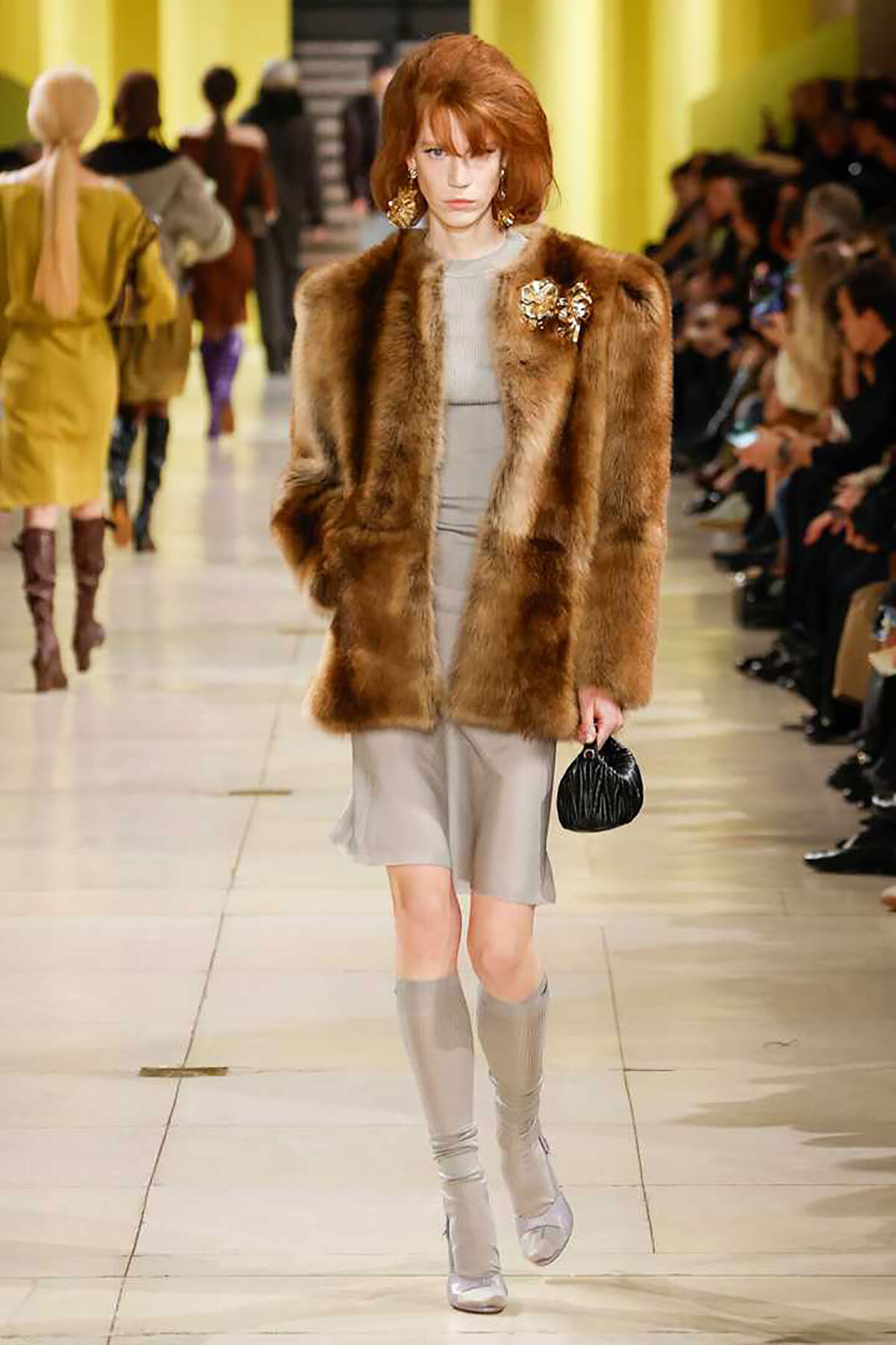 Runway model wears a brown fur coat over a gray dress, paired with taupe knee-high boots and a small black handbag, during a fashion show theme with yellow backdrop.