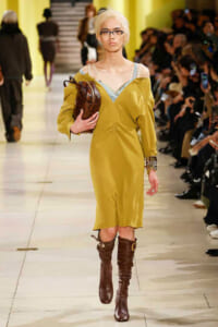 Model walking a fashion runway in a mustard-yellow off-shoulder dress, blue undergarment straps visible, brown knee-high boots, and a brown handbag; audience on both sides.