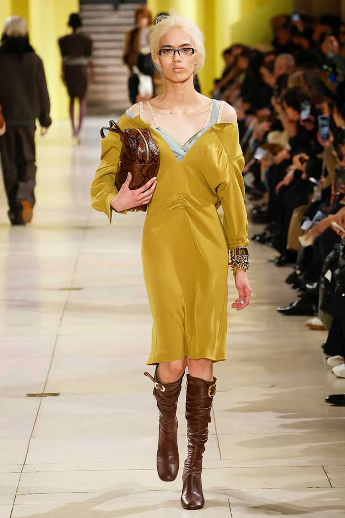 Model walking a fashion runway in a mustard-yellow off-shoulder dress, blue undergarment straps visible, brown knee-high boots, and a brown handbag; audience on both sides.