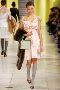 Model walking runway in pale pink satin dress, carrying a light green handbag and a brown fur stole over her arm.