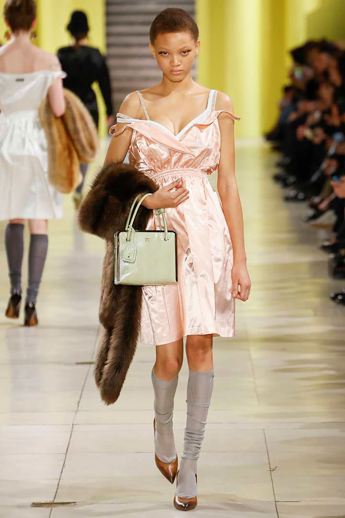 Model walking runway in pale pink satin dress, carrying a light green handbag and a brown fur stole over her arm.