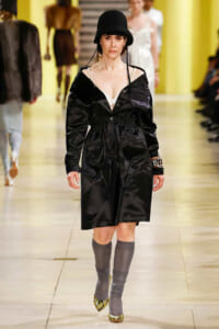 Model walking a runway in a black belted coat, white top, gray knee-high socks, gold shoes, and a black bucket hat.