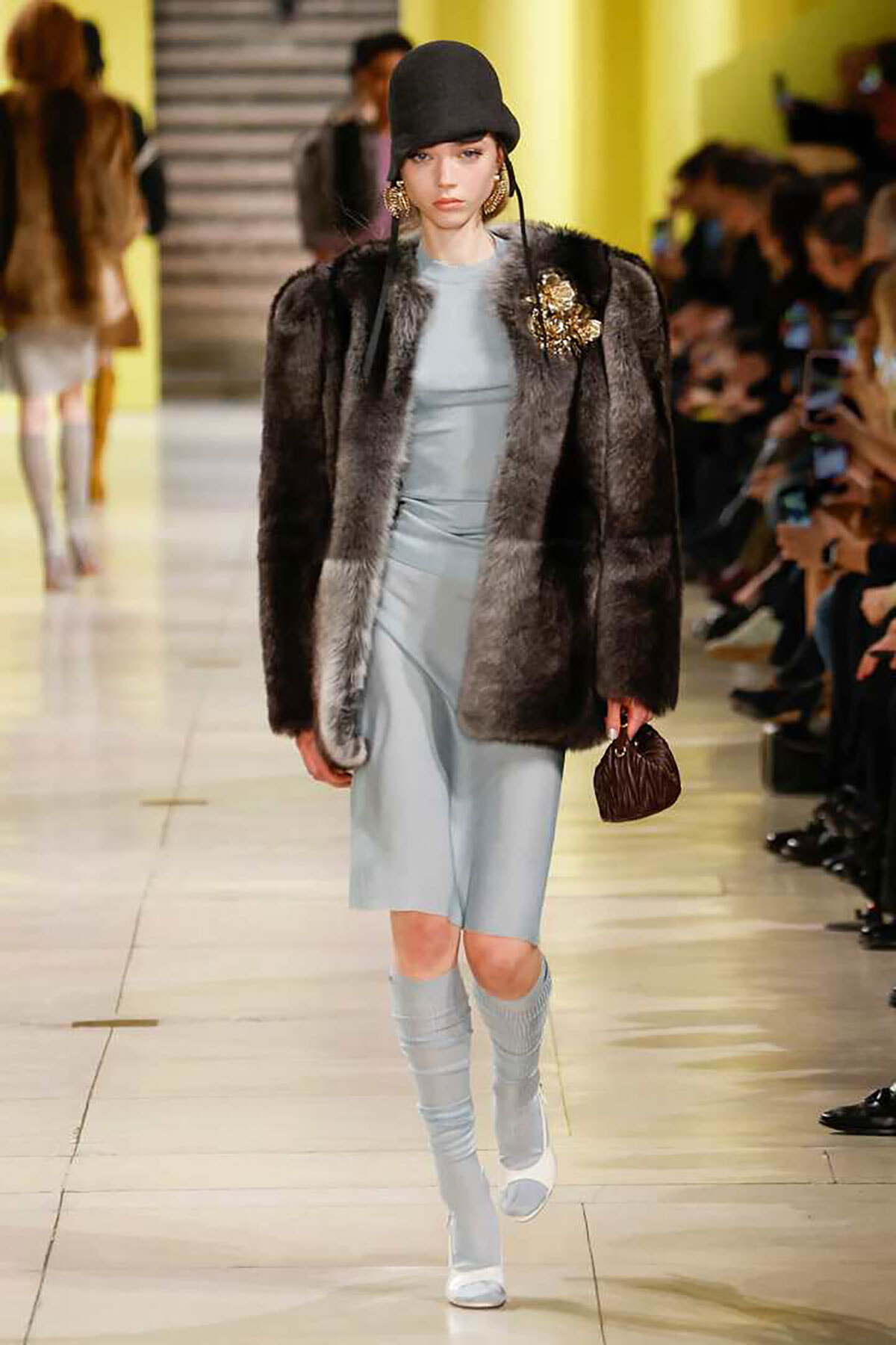 Model walks the runway in a gray-blue satin dress under a dark fur coat, with a black hat, gold earrings, and a small brown handbag.