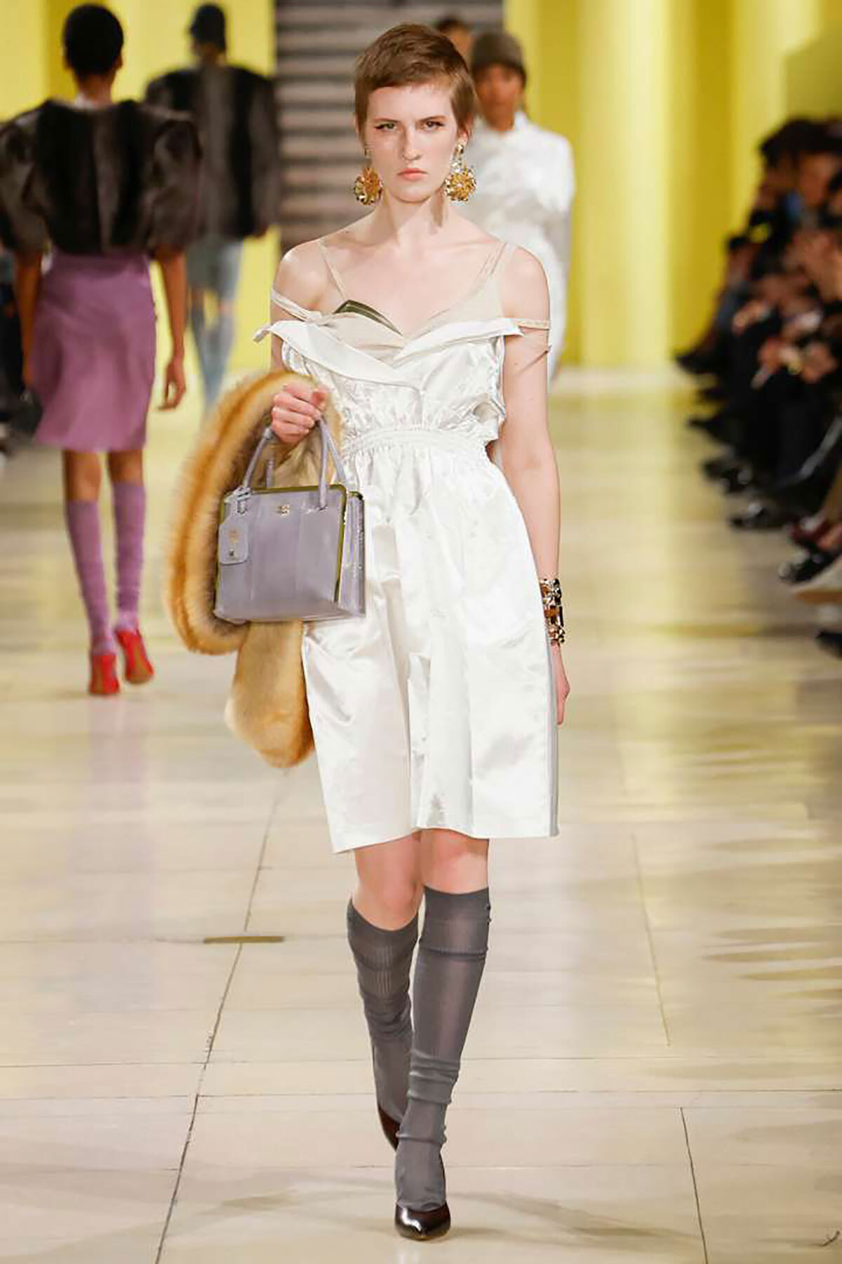 Model on a fashion runway wearing a white off-shoulder dress, holding a lavender handbag and fur stole, with grey knee-high boots.