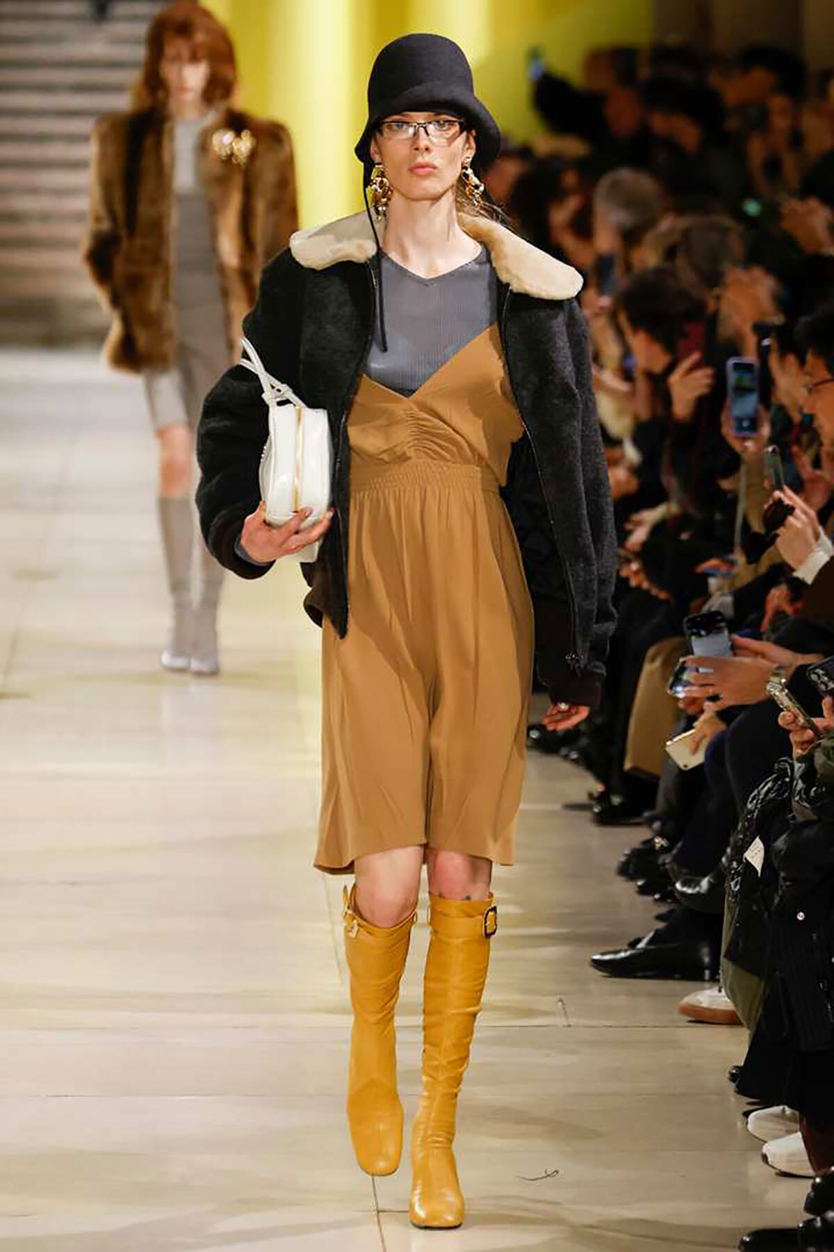 Model walks a fashion runway in a tan jumpsuit over a grey top, black shearling jacket, and mustard knee-high boots, wearing a black hat and glasses while carrying a white bag.
