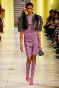 Model walking the runway in a lavender sheath dress paired with a gray fur stole, lavender knee-high socks, red heels, and a small gray handbag.
