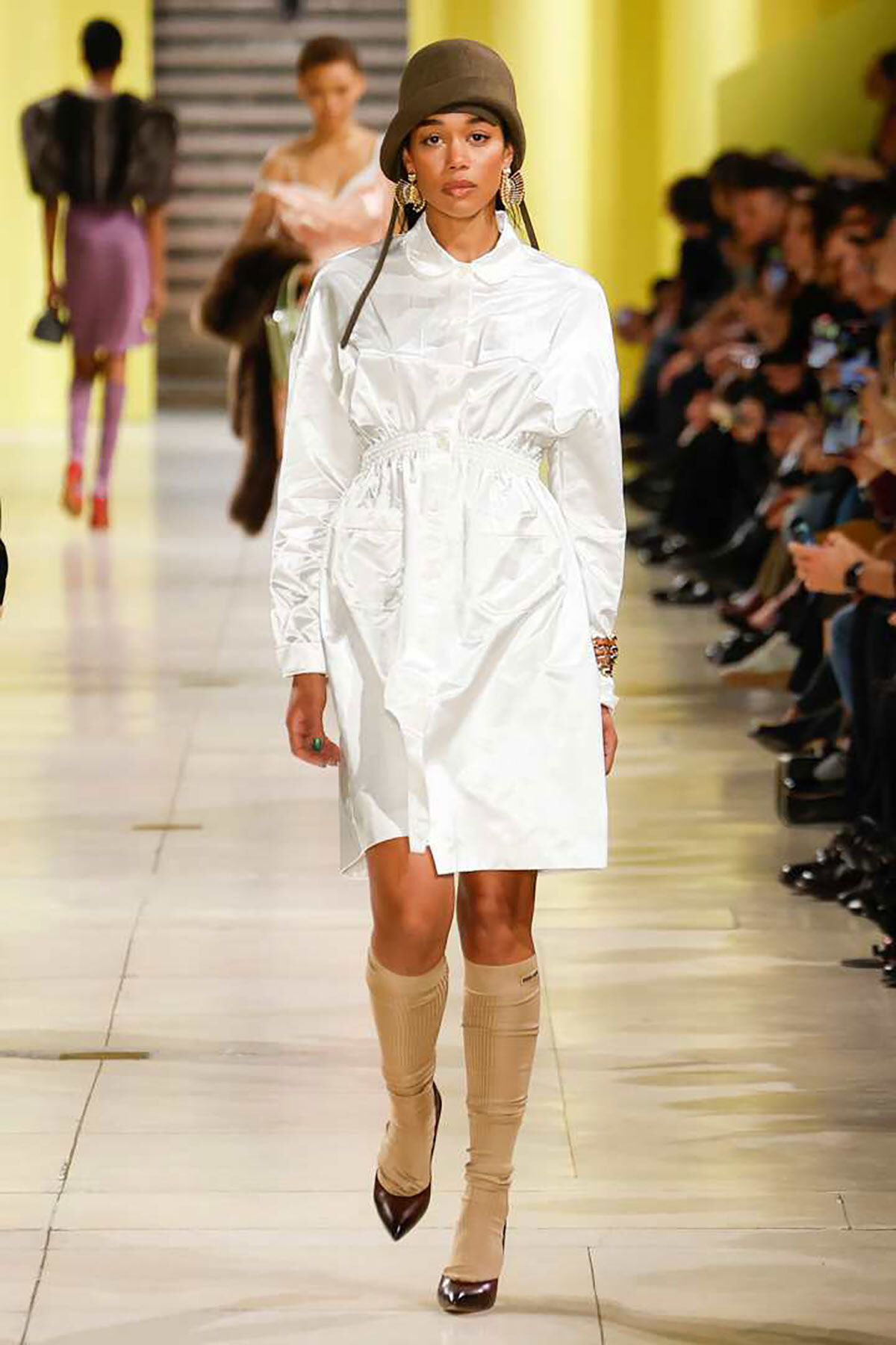 Model walks the runway in a white belted shirt dress, beige knee-high boots, and a brown bucket hat with large earrings.